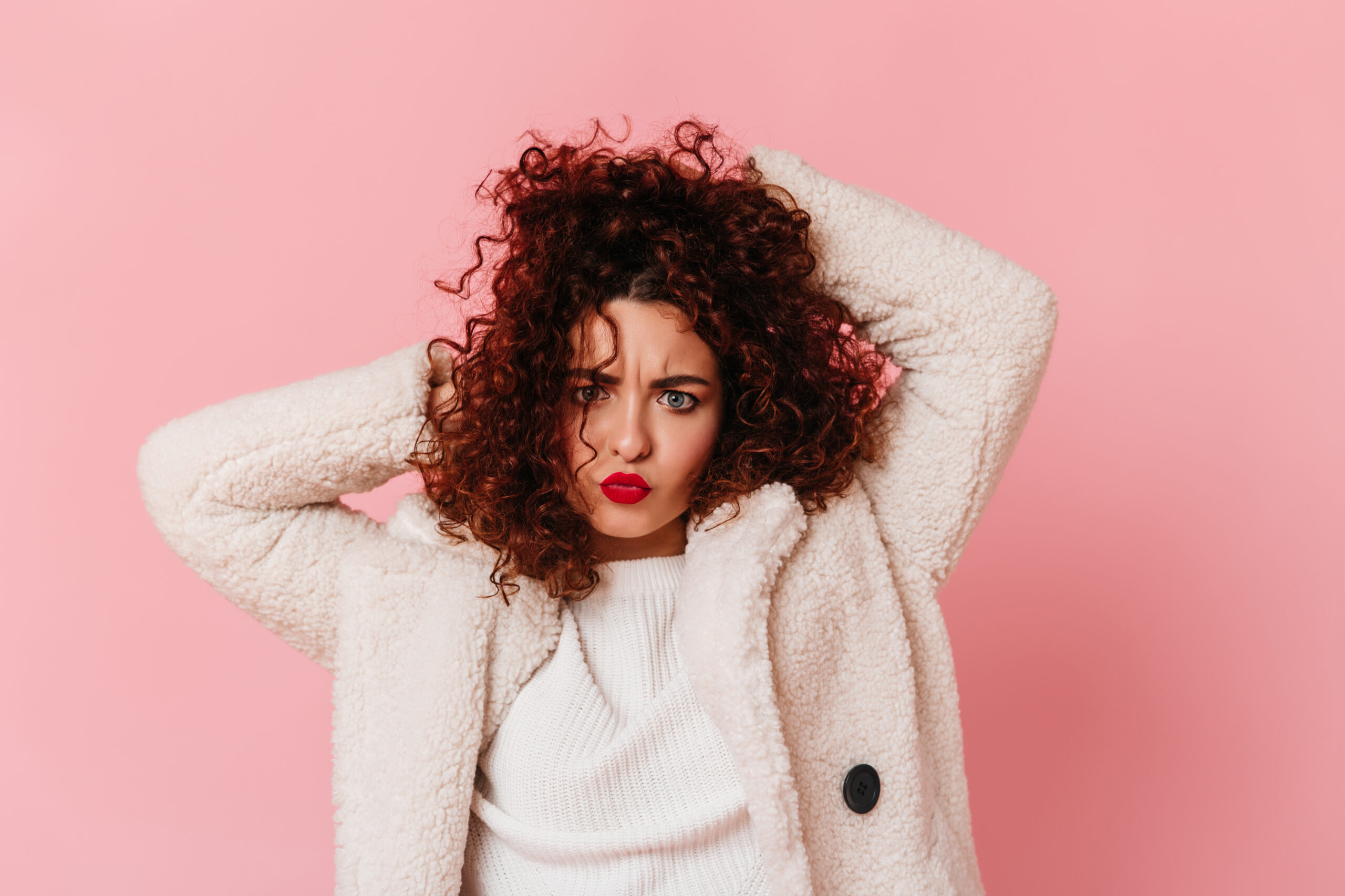 Cheerful girl in white warm outfit tousles her curly dark hair and looks into camera on pink backgr